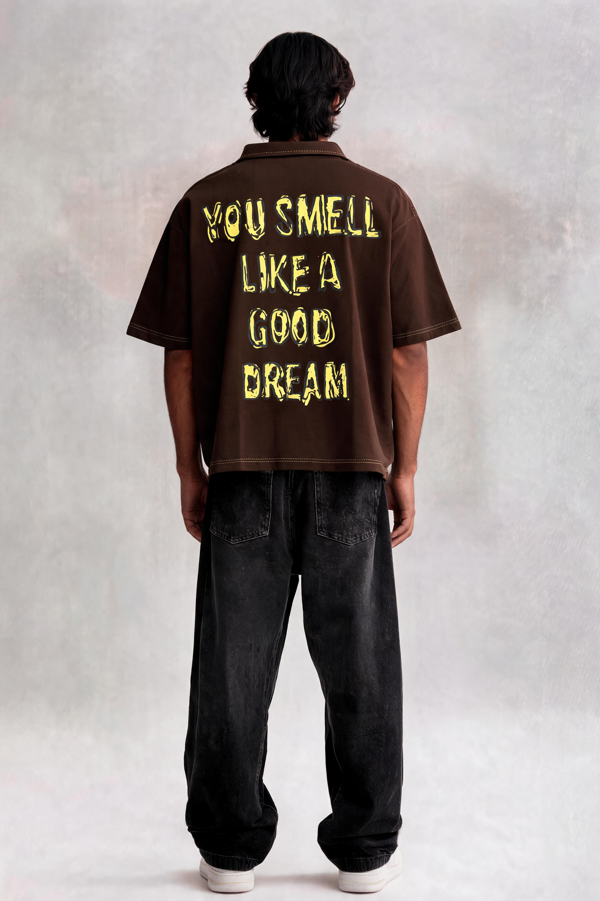 Person wearing a brown t-shirt with yellow text on the back, standing against a plain background