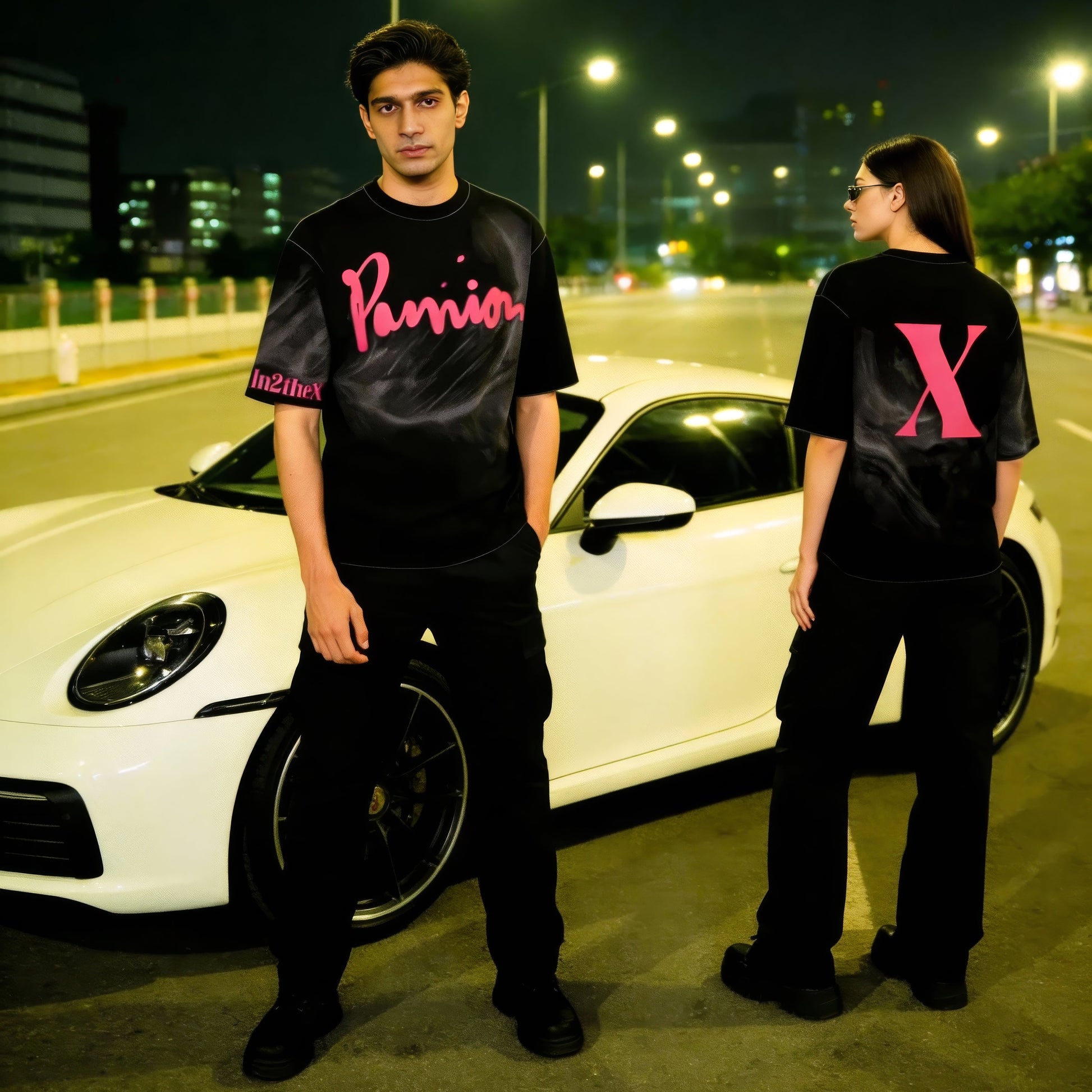 Two people wearing black t-shirts with pink logos standing next to a white car on a street at night.